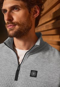 Felpa leggera grigia con zip a quarto di pollice e pannello superiore texture, zip nera e patch con logo sul petto. Tessuto morbido con colletto a coste.