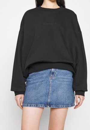 Kobieta ubrana w czarną bluzę Adidas i niebieską mini spódniczkę dżinsową, stojąca na jednolitym białym tle.