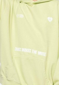 Sudadera color verde claro hecha de tejido suave, con un bolsillo canguro, texto blanco "EL CAOS INSPIRA A LA MUSA" y acentos de logo en el pecho.
