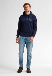 Námořnicky modrá mikina s kapucí a šňůrkami, potiskem loga, doplněná světle modrými džínami a tmavě hnědými botami. Hladká textura a pohodlný střih.