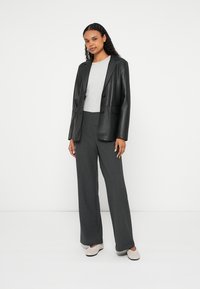 Melns ādas žakete, pelēks džemperis un platās pelēkās bikses. Modelis stāv pārliecinoši, valkājot bēšas slip-on kurpes.