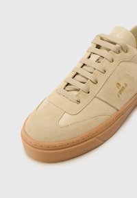 Sneaker in suede e pelle beige con punta rotonda, suola in gomma e lacci. Presenta dettagli testurizzati e un piccolo logo dorato sul lato.