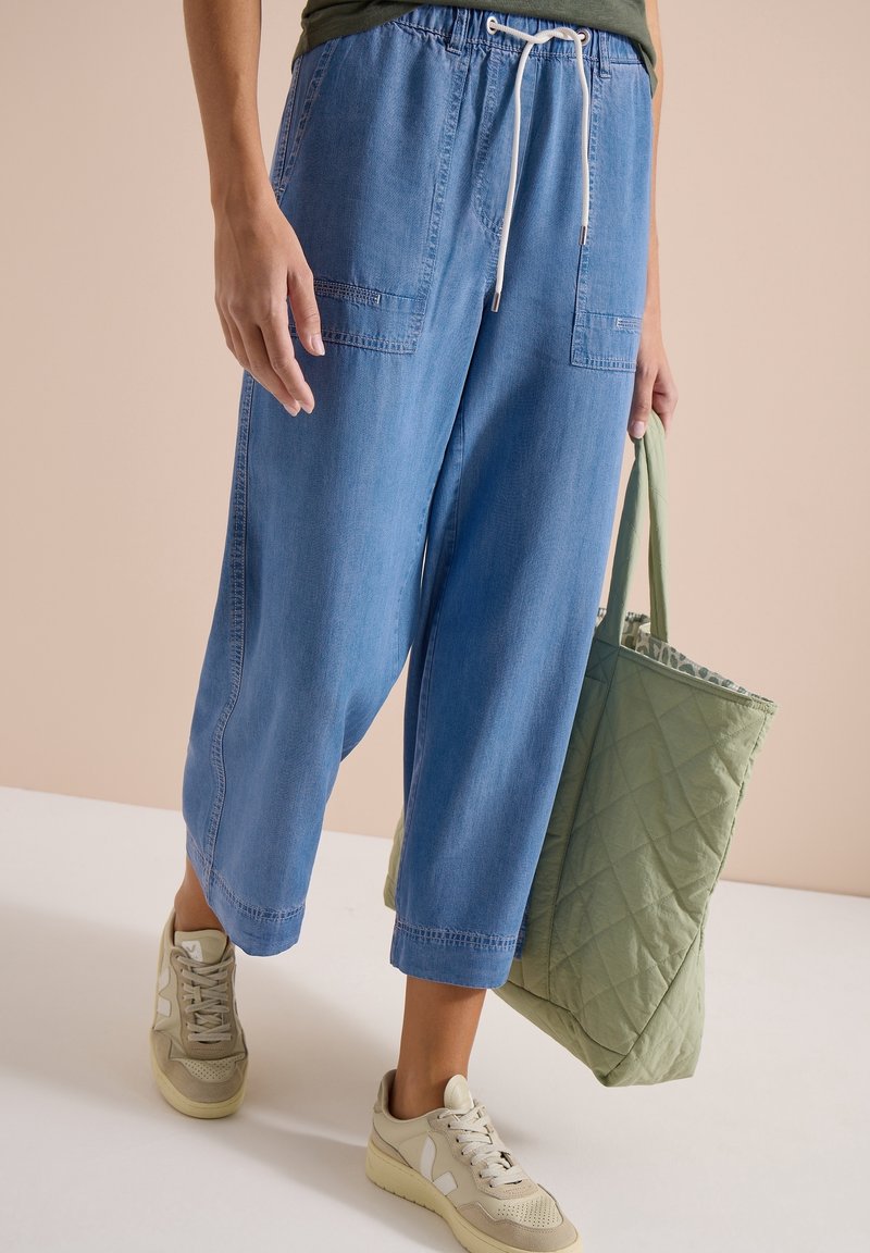 Personne portant un pantalon en jean bleu ample et des baskets beiges tenant un grand sac cabas matelassé vert à ses côtés.
