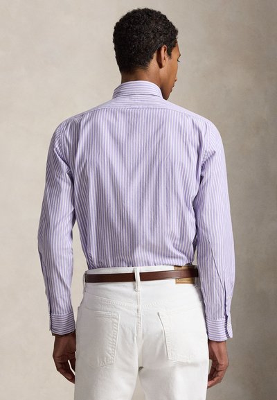 Άνδρας που φοράει μακρυμάνικο πουκάμισο με ριγέ μοβ και λευκό, κρυμμένο μέσα σε λευκό παντελόνι με καφέ ζώνη, από πίσω.