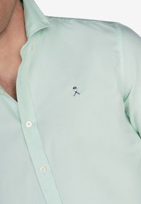 Camisa de botón verde menta, fabricada en algodón, con un pequeño logo bordado. Textura suave, cuello clásico y cierre de botón estándar.