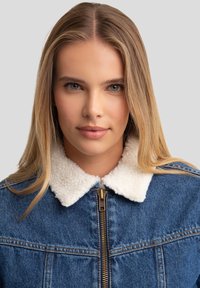 Džinsinė striukė su šviesios spalvos, tekstūruotu dirbtinio pūkų apykakle. Turi priekinį užtrauktuką ir kontrastingą siuvinėjimą.