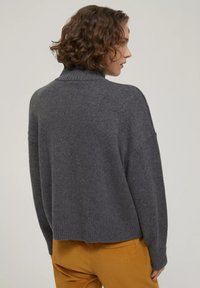 Grauer Strickpullover mit hohem Kragen, überschnittenen Schultern und geripptem Saum, kombiniert mit senfgelben Cordhosen. Weiche Textur, lockere Passform.