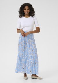 Jupe maxi florale bleu clair avec une taille smockée, une conception superposée et une texture fluide. Associée à une chemise blanche ajustée et des ballerines beiges.