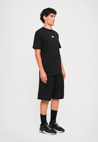 Νεαρός άνδρας που στέκεται φορώντας μαύρο φαρδύ μπλουζάκι, μαύρες σορτσάτες παντελόνες μέχρι το γόνατο, μαύρες κάλτσες και μαύρα αθλητικά παπούτσια με λευκή σόλα.