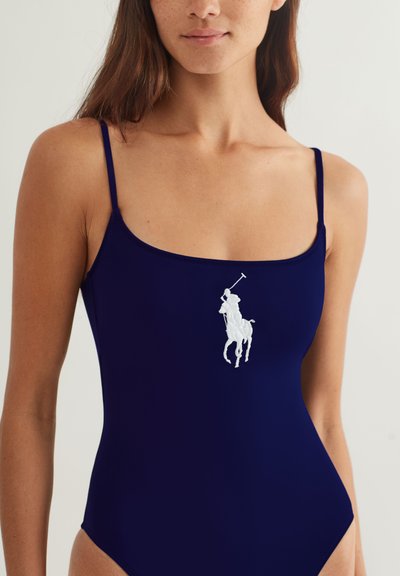 Γυναίκα που φορά μπλε ναυτικό αμάνικο τοπ με λεπτούς ιμάντες και λευκό λογότυπο παίκτη πόλο στο κέντρο του στήθους, μπροστά σε απλό φόντο.