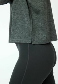 Nahaufnahme des Torsos, der ein dunkelgraues, langärmeliges Oberteil mit seitlichem Schlitz und schwarze, enganliegende Leggings mit sichtbaren Nähten trägt.