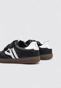 Zapatillas negras y blancas con acabado de cuero liso, que cuentan con una cinta de velcro, suela de goma y costuras decorativas en blanco.