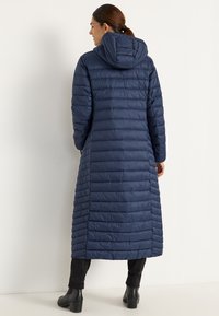 Marinblå isolerad lång jacka med horisontellt quiltat mönster, huva och sidoflikar, tillverkad av glänsande, slätt material, bärs över svarta byxor.