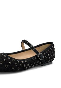 Zapato plano negro de ante con punta redonda, correa en el tobillo y tachuelas decorativas a lo largo de los lados. Forro interior suave y tacón bajo.