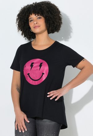 Frau mit lockigem Haar, die ein schwarzes T-Shirt mit einem pinken Smiley mit Blitzaugen trägt, steht mit einer Hand in der Hüfte.