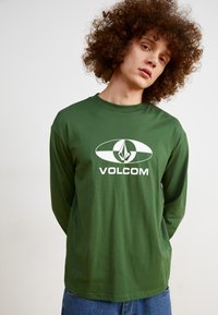 Langermet grønn skjorte med en hvit Volcom-logo. Laget av et glatt stoff, har den rund hals og en avslappet passform.