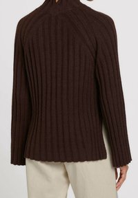 Brązowy sweter z długim rękawem o prążkowanej fakturze noszony przez osobę, której widoczna jest jedna ręka, zestawiony z jasnoszarymi spodniami na jednolitym tle.