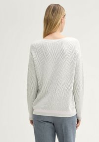 Lichtgrijze trui met een visgraatpatroon, bootnek en geribbelde zoom. Gepaard met grijze broek, die een ontspannen pasvorm laat zien.