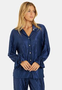 Camisa de satén azul marino con un patrón ondulado texturizado, mangas largas con puños atados y detalles de botón en la parte delantera. Acabado suave y corte relajado.
