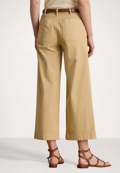 Pantalon beige à jambes larges avec une taille haute, comportant deux poches arrière, associé à des sandales brunes à lanières. Texture en coton lisse, design décontracté.