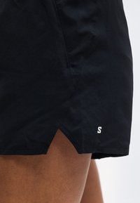 Κοντινό πλάνο μαύρων αθλητικών σορτς με πλαϊνή σχισμή και μικρό λευκό λογότυπο "S" στο κάτω στρίφωμα, φορεμένα στον πάνω μηρό ενός ατόμου.