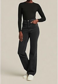 Personne portant un haut noir ajusté à manches longues, un jean noir taille haute à jambes larges, et des chaussures blanches sans lacets, debout avec une main dans la poche.