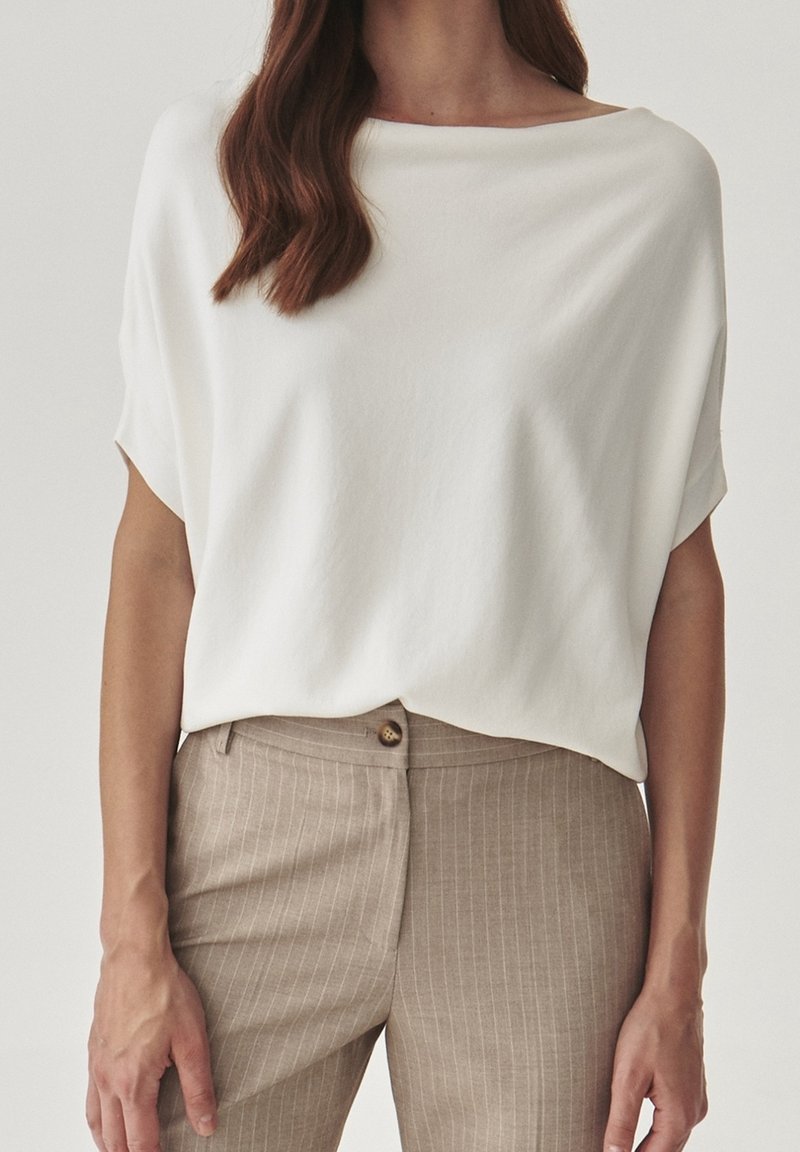 Femme portant un chemisier blanc ample à manches courtes, rentré dans un pantalon taille haute beige à fines rayures avec une fermeture à boutons.