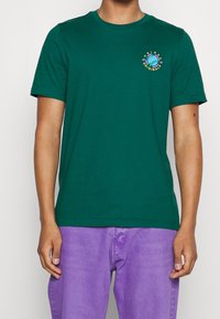 Muž v zeleném tričku Adidas Originals s malým logem globu na hrudi, v kombinaci s fialovými kalhotami, stojící na bílém pozadí.