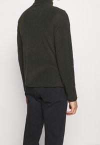 Dark green fleece jacket with high collar, zippered closure, and long sleeves, paired with black pants. Smooth texture, minimal design.