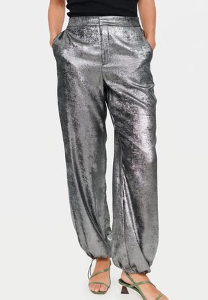 Glänzende silberne locker sitzende Hose mit elastischen Bündchen, getragen mit grünen Riemchenabsätzen und einem schwarzen ärmellosen Top, Hände in den Taschen.