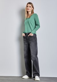 Grünes langärmliges Hemd kombiniert mit schwarzen weiten Jeans, die über Fronttaschen verfügen, und weißen Sneakers vor einem grauen Hintergrund.