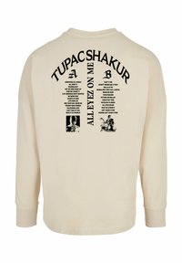 Cămașă bej cu mâneci lungi, având text și imagini imprimate în negru pe spate, inclusiv titluri de melodii și numele artistului "TUPAC SHAKUR."