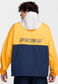 Chaqueta cortavientos amarilla y azul marino con capucha, con la palabra "SPORTSWEAR" impresa en la parte posterior, fabricada con un material suave y ligero.