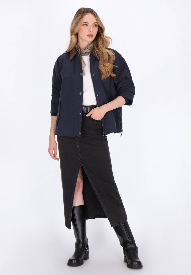 Marineblau Bomberjacke, weißes T-Shirt, schwarzer Jeansrock mit Schlitz vorne und schwarze Knöchelstiefel. Das Model trägt einen beigen gemusterten Schal.