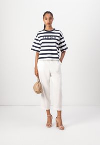 Marineblå og hvide stribede bomulds-t-shirt med korte ærmer og teksten "STUDIO", parret med hvide linne cropped bukser og en beige håndtaske.