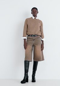 Un pull en maille côtelée marron porté par-dessus une chemise blanche à col, associé à des culottes en denim beiges et des bottes noires à talons hauts jusqu'aux genoux.