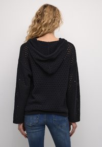 Melnas, ar kapuci, džemperis ar atvērtu adījumu. Dizainā ir garas piedurknes un brīvs griezums, kā arī ribota apakšmala un piedurkņu gali.