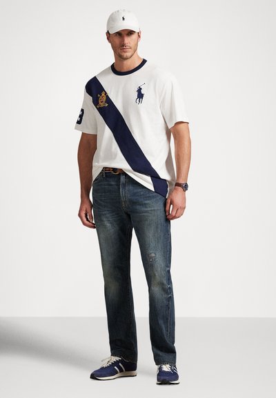 Λευκό κοντομάνικο T-shirt με ναυτικό διαγώνιο ρίγα, διακοσμημένο με λογότυπο και αριθμό. Φοριέται με μπλε τζιν με φθορές και ναυτικά αθλητικά παπούτσια.