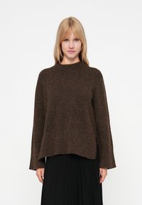 Sweater castanho trançado com gola alta, de corte descontraído e textura canelada. Combinado com uma saia preta plissada, mostrando um visual em camadas.