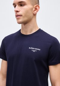 Νέος άνδρας με κοντά μαλλιά που φοράει μπλε σκούρο μπλουζάκι Björn Borg με λευκή λογότυπο στο στήθος, στραμμένος ελαφρώς προς τα αριστερά.