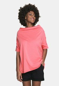 Koronkowy, oversize'owy t-shirt w kolorze koralowym z szerokim dekoltem i krótkimi rękawami, wykonany z miękkiego materiału, w połączeniu z czarnymi szortami. Prosty design.