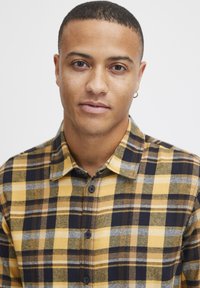 Kollane ja must ruuduline flanellshirt hallide detailidega, varustatud nööbitava kraega ja esise konstruktsiooniga, valmistatud pehmest kangast.