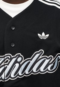 Μαύρο ποδοσφαιρικό τζέρσεϊ με λευκό γιακά και κεντημένο κείμενο. Διαθέτει τρεις λευκές ρίγες στους ώμους και ένα μικρό λευκό λογότυπο.