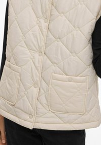 Beežitud tikandiga vest rombimustriga, krõpsu nööpidega ja kahe eesmise taskuga; kangas tundub pehme ja veidi läikiv.