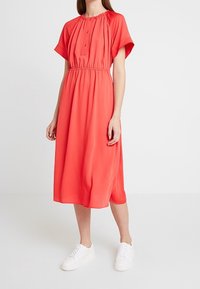 Robe midi corail avec un décolleté froncé et des manches courtes ; elle possède une taille élastique et des détails de boutons, associée à des baskets blanches.