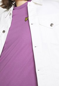 Biała dżinsowa kurtka na purpurowym t-shircie z żółtym haftowanym motylem w pobliżu kołnierza, charakteryzująca się gładką fakturą i prostym wzornictwem.