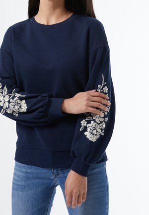 Personne portant un pull bleu marine avec une broderie florale blanche sur les manches bouffantes et un jean bleu, sur un fond blanc uni.
