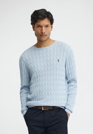 Uomo con capelli scuri che indossa un maglione azzurro chiaro a trecce e pantaloni scuri con le mani nelle tasche, sfondo neutro.