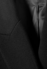 Κοντινό πλάνο του πέτου ενός μαύρου παλτού από μαλλί πάνω από το υφασμάτινο γιακά ενός γκρι πουκάμισου με υφή, αναδεικνύοντας τις λεπτομέρειες του υφάσματος και τις ραφές.