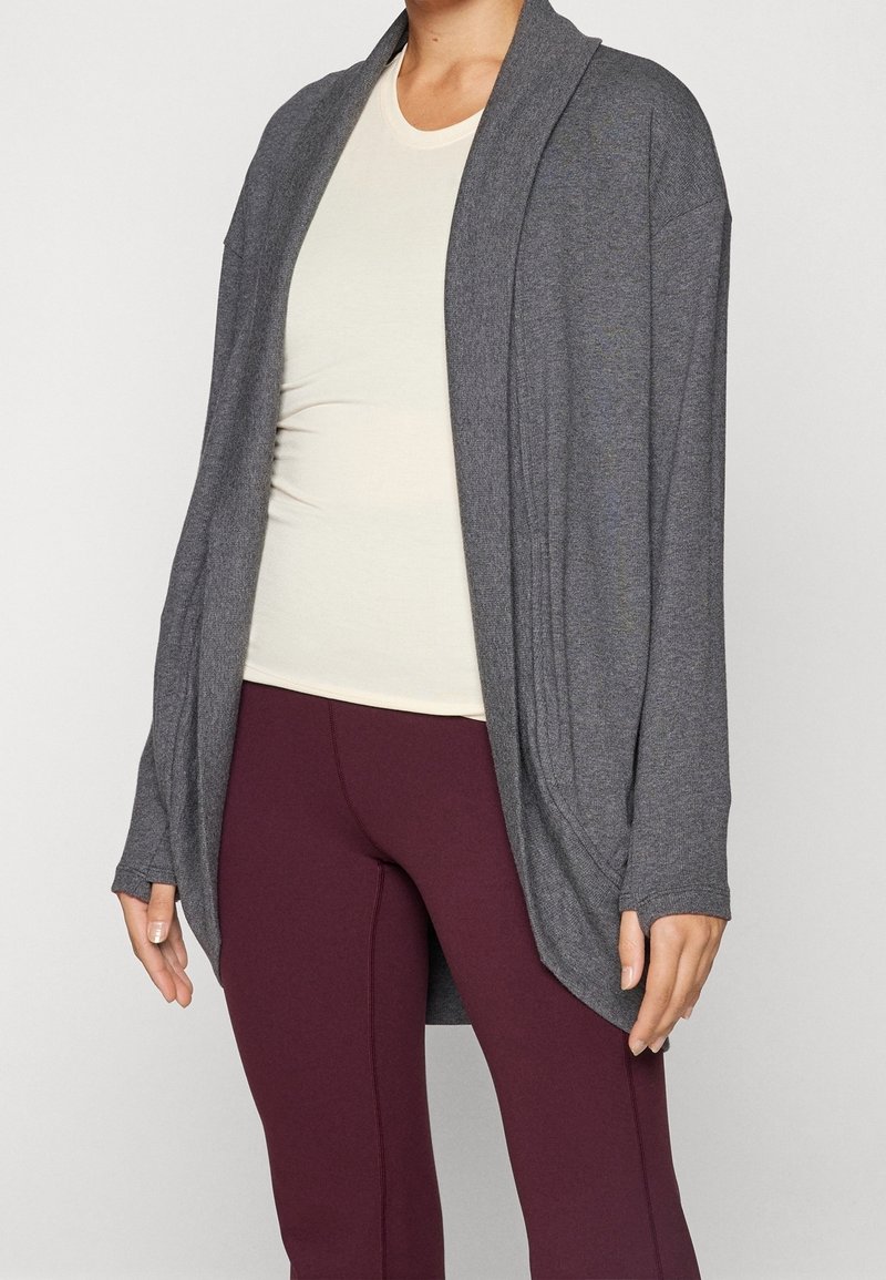 Personne portant un haut ajusté beige clair, des leggings bordeaux et un cardigan ample gris foncé, ouvert sur le devant, avec des manches longues.